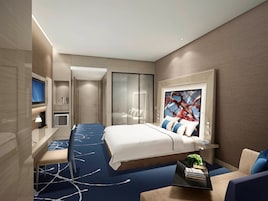 Una habitación de hotel moderna con una cama grande, un escritorio con televisor de pantalla plana, una zona de estar con sofá y sillones, y un cuadro abstracto de gran tamaño en la pared.