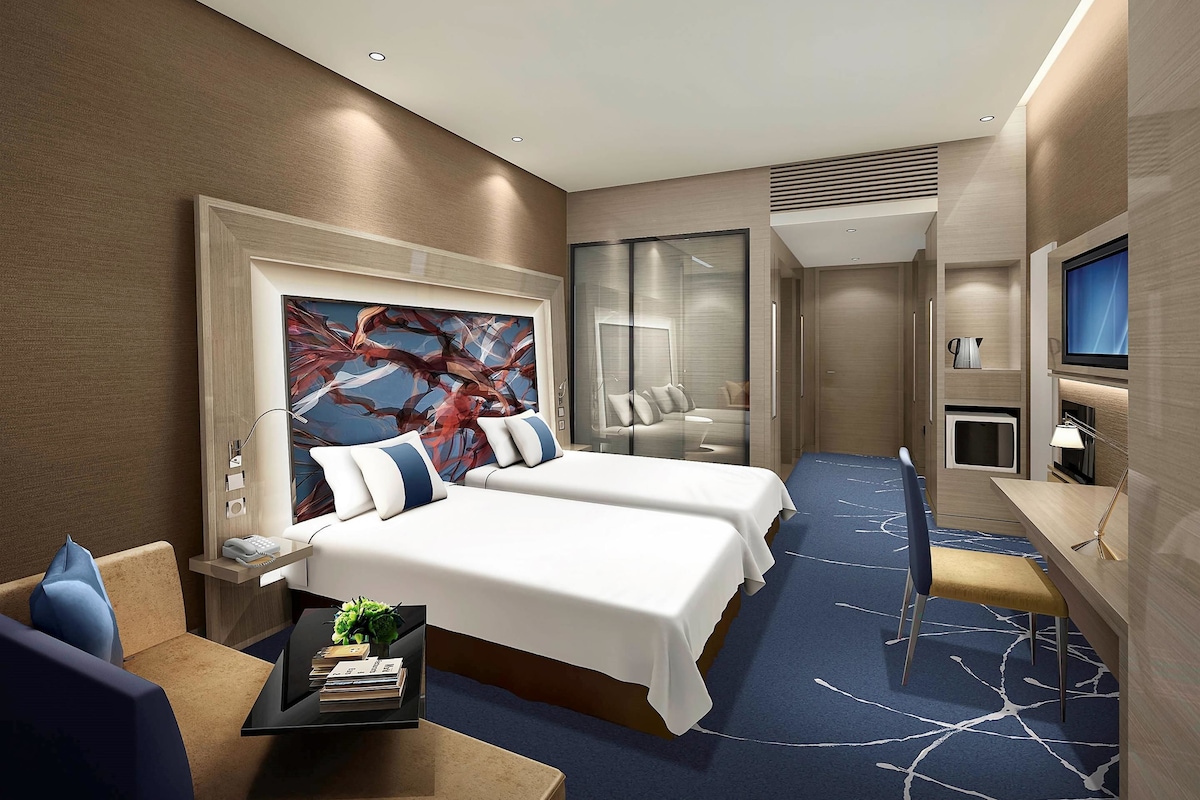 Habitación de hotel con dos camas, un escritorio, un televisor y un cuadro abstracto grande en la pared.