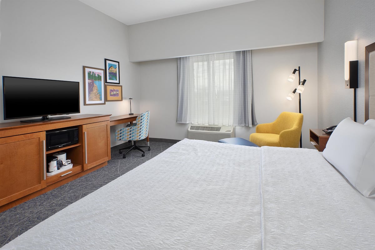 Una camera d'albergo con un letto, una scrivania con una TV a schermo piatto, una poltrona gialla, una lampada e quadri appesi al muro.