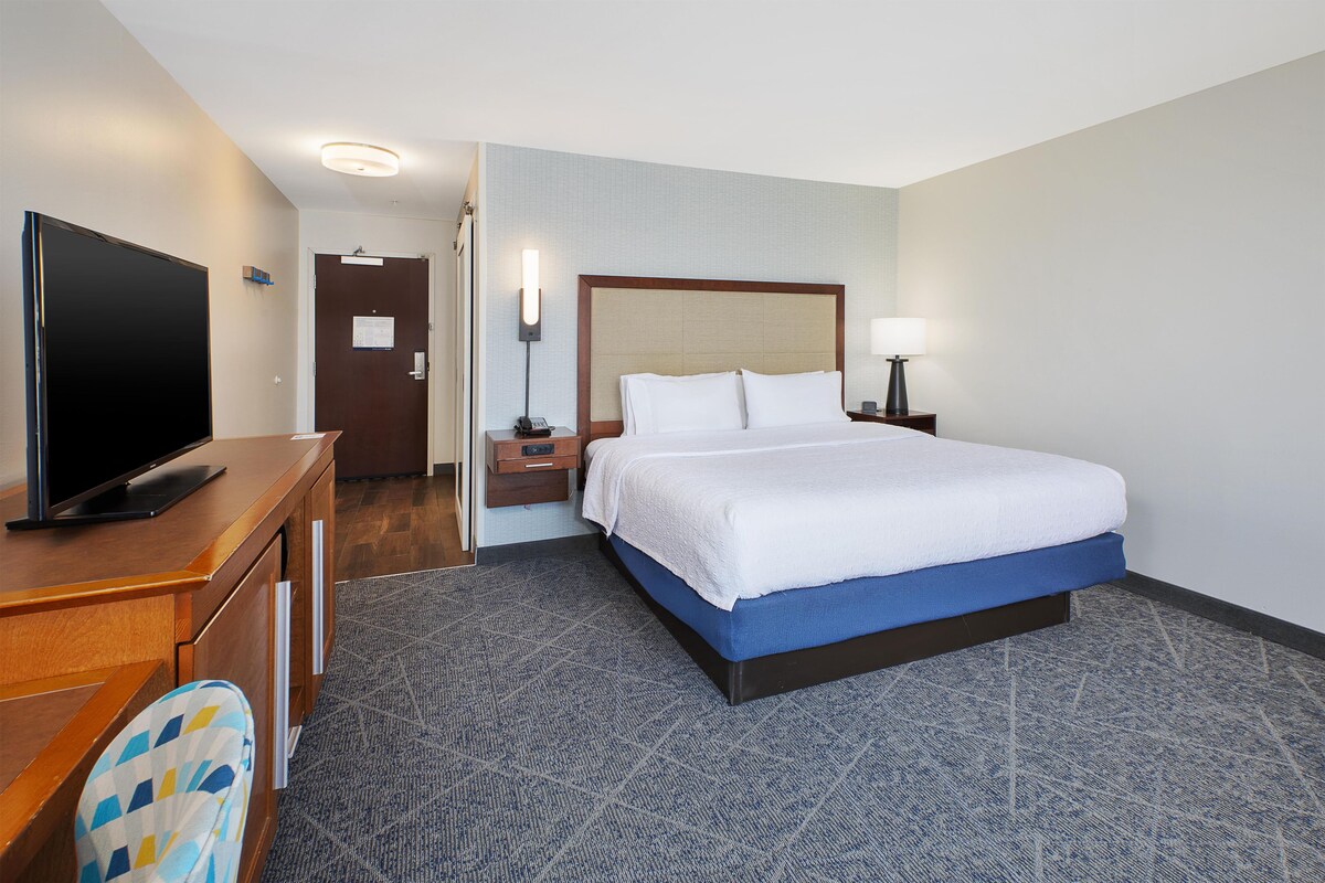 Une chambre d’hôtel comprenant un lit, un bureau avec un téléviseur à écran plat, une chaise, une table de chevet avec une lampe et une porte avec une pancarte.