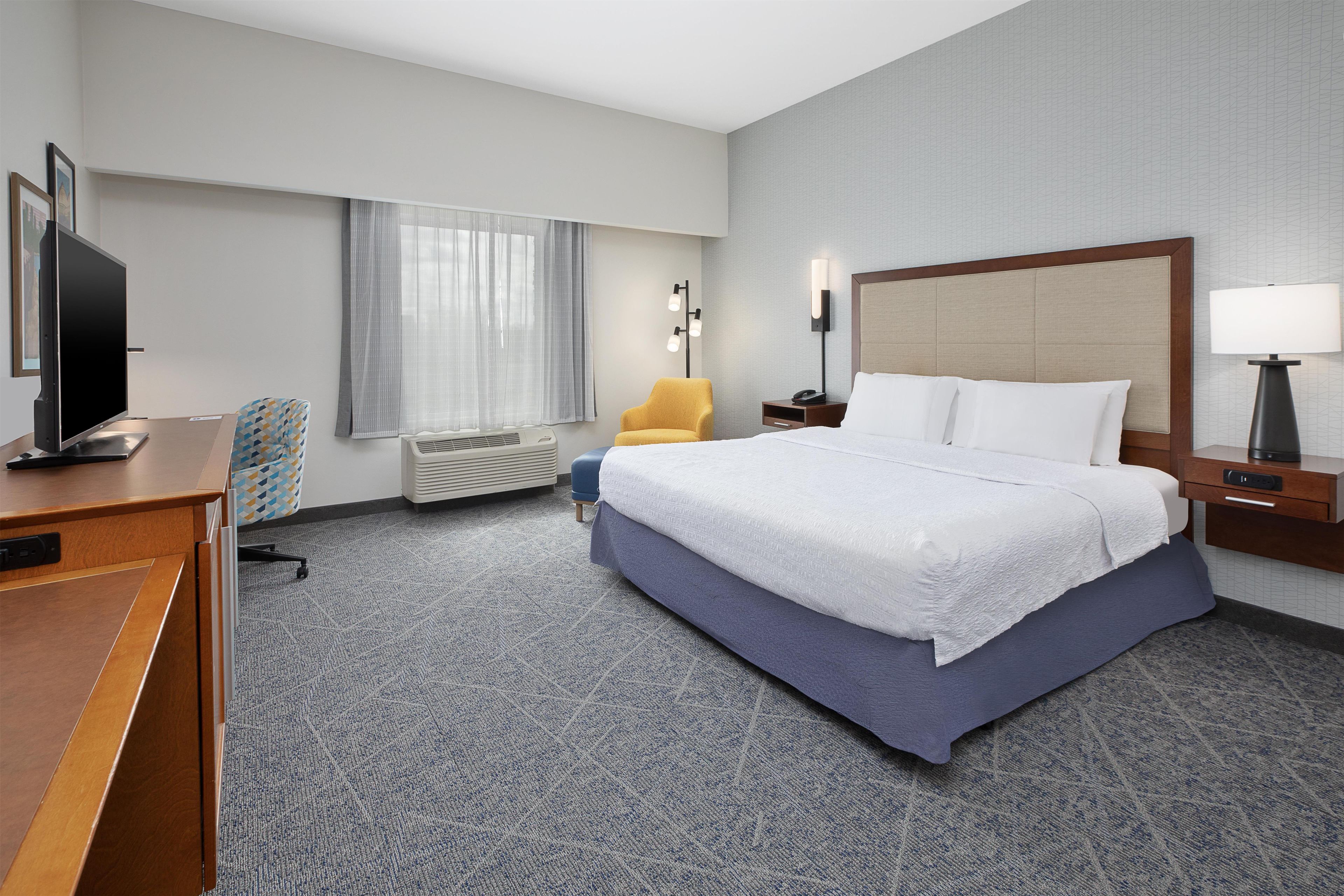 A hotel room with a bed, desk, TV, chair, lamp, and window with curtains.