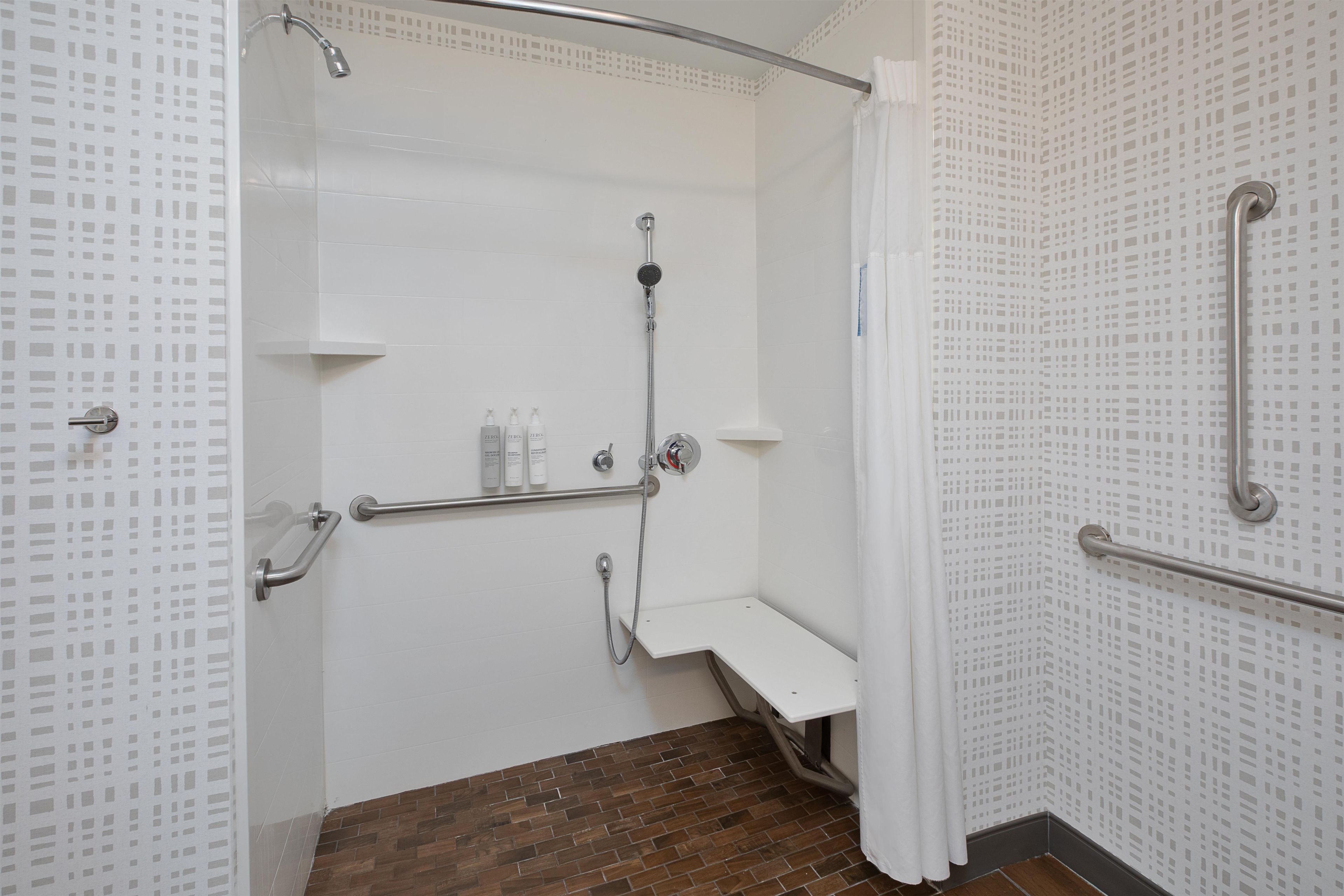Un espace douche avec un mur recouvert de carreaux blancs, un pommeau de douche, un pommeau de douche à main, un banc et une barre d’appui.