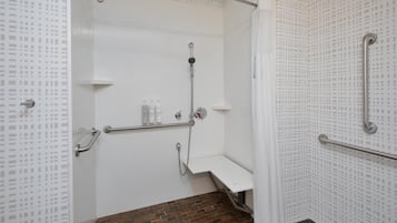 Un espace douche avec un mur recouvert de carreaux blancs, un pommeau de douche, un pommeau de douche à main, un banc et une barre d’appui.