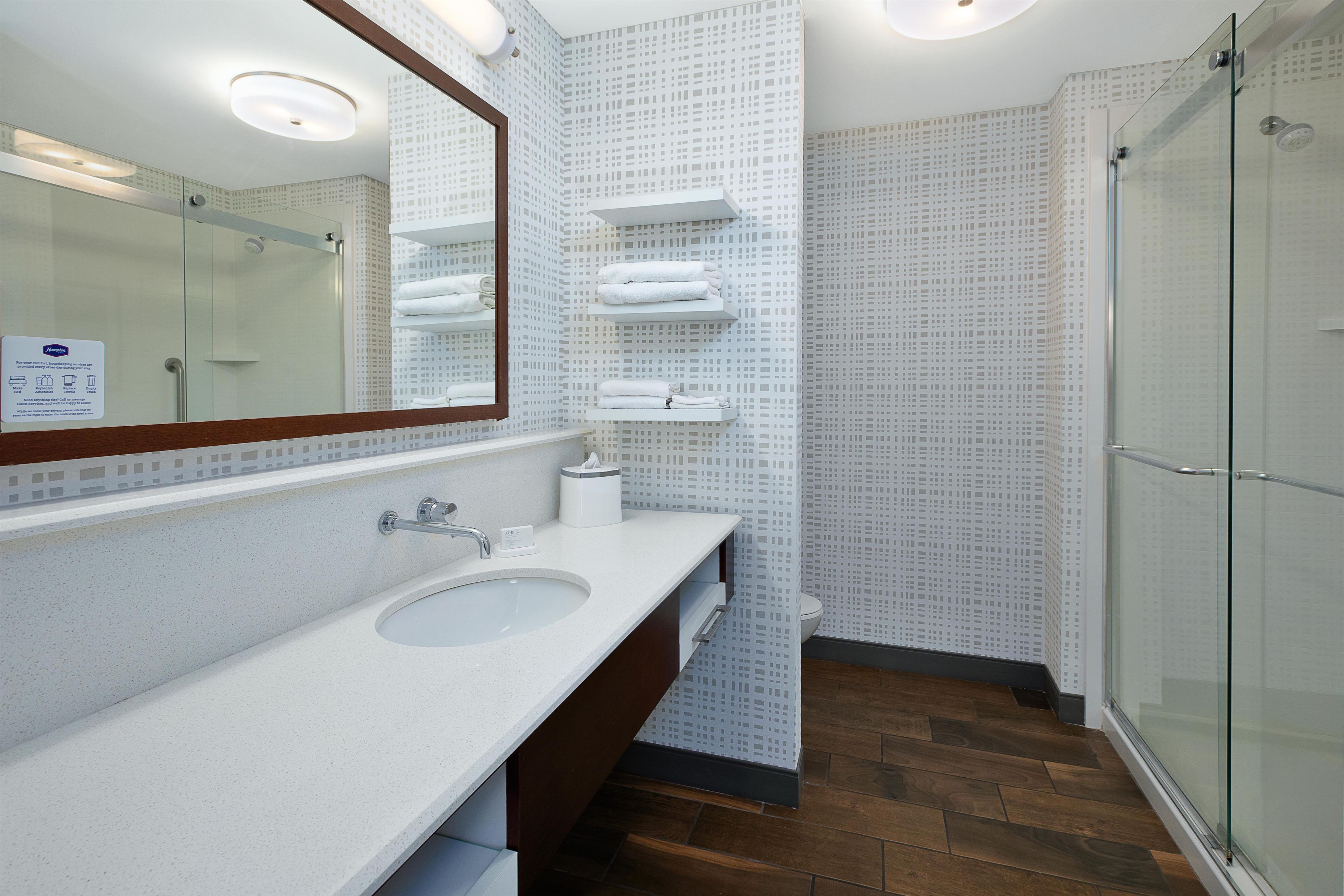 Un baño con lavamanos, espejo, toalleros y mampara de cristal.