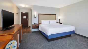Ein Hotelzimmer mit einem Bett, einem Schreibtisch mit Flachbildfernseher, einem Sessel, einem Nachttisch mit Lampe und einer Tür mit Hinweisschild.