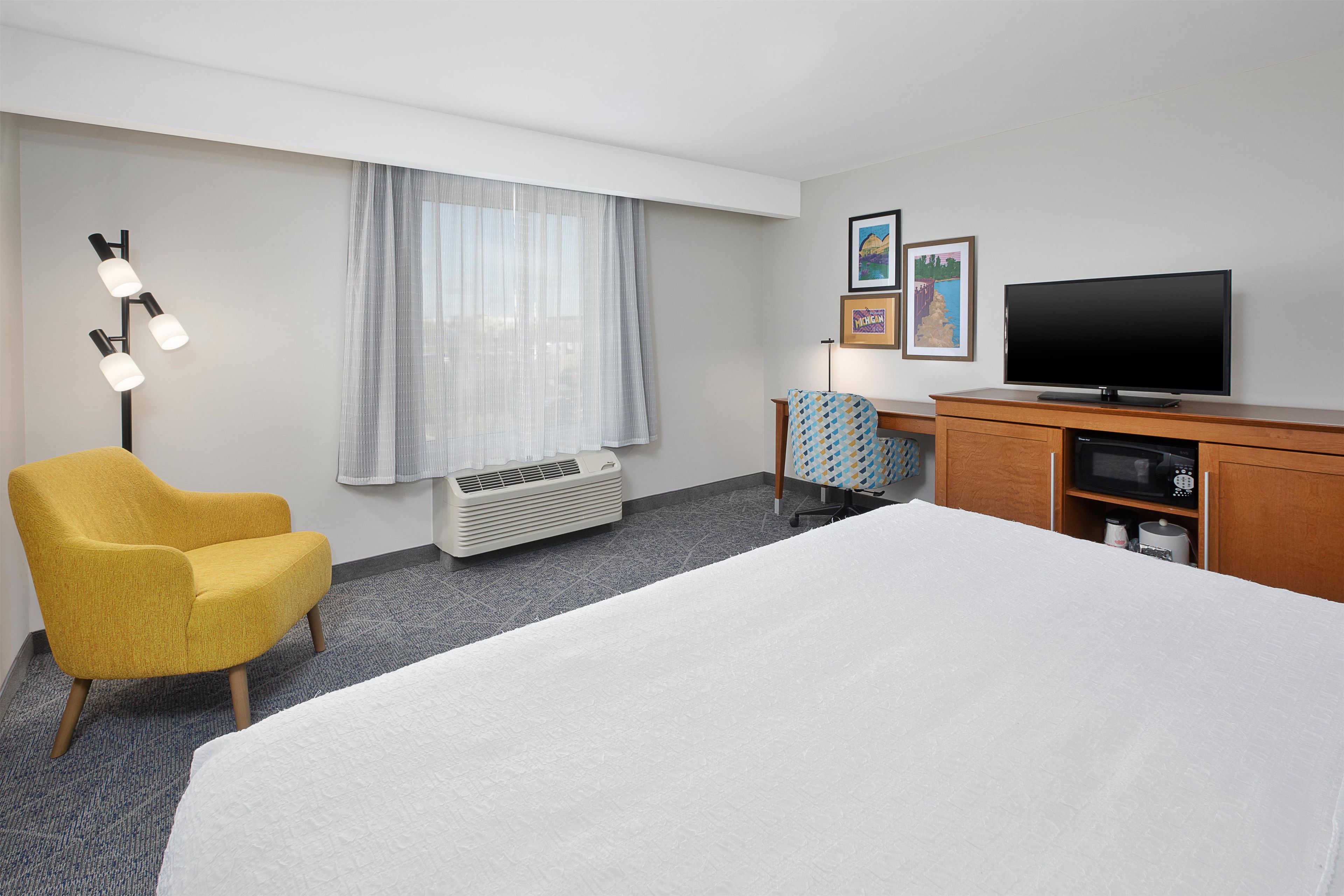 Ein Hotelzimmer mit einem Bett, einem gelben Sessel, einem Schreibtisch mit Fernseher und einem Fenster mit Vorhängen.