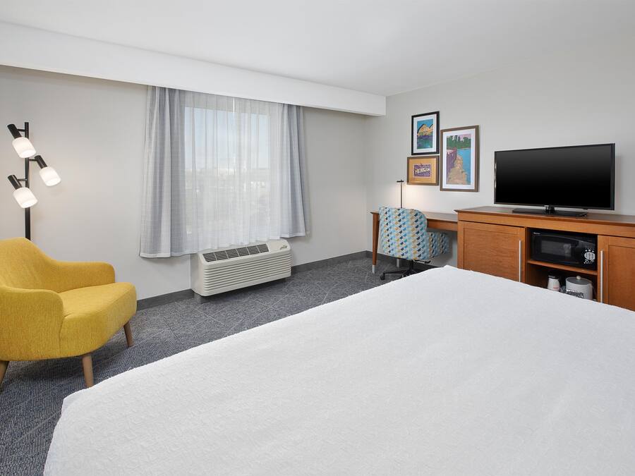 Ein Hotelzimmer mit einem Bett, einem gelben Sessel, einem Schreibtisch mit Fernseher und einem Fenster mit Vorhängen.