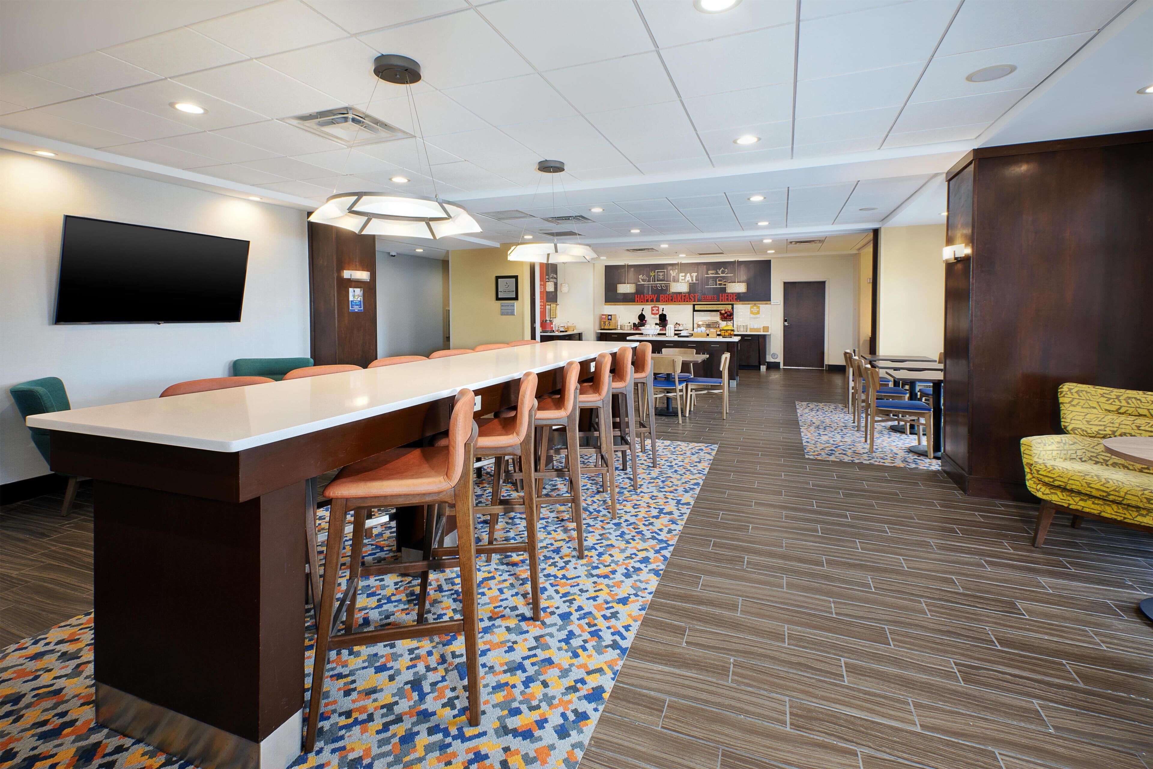 A hotel room with a long dining table, multiple chairs, a kitchen area, and a TV mounted on the wall.