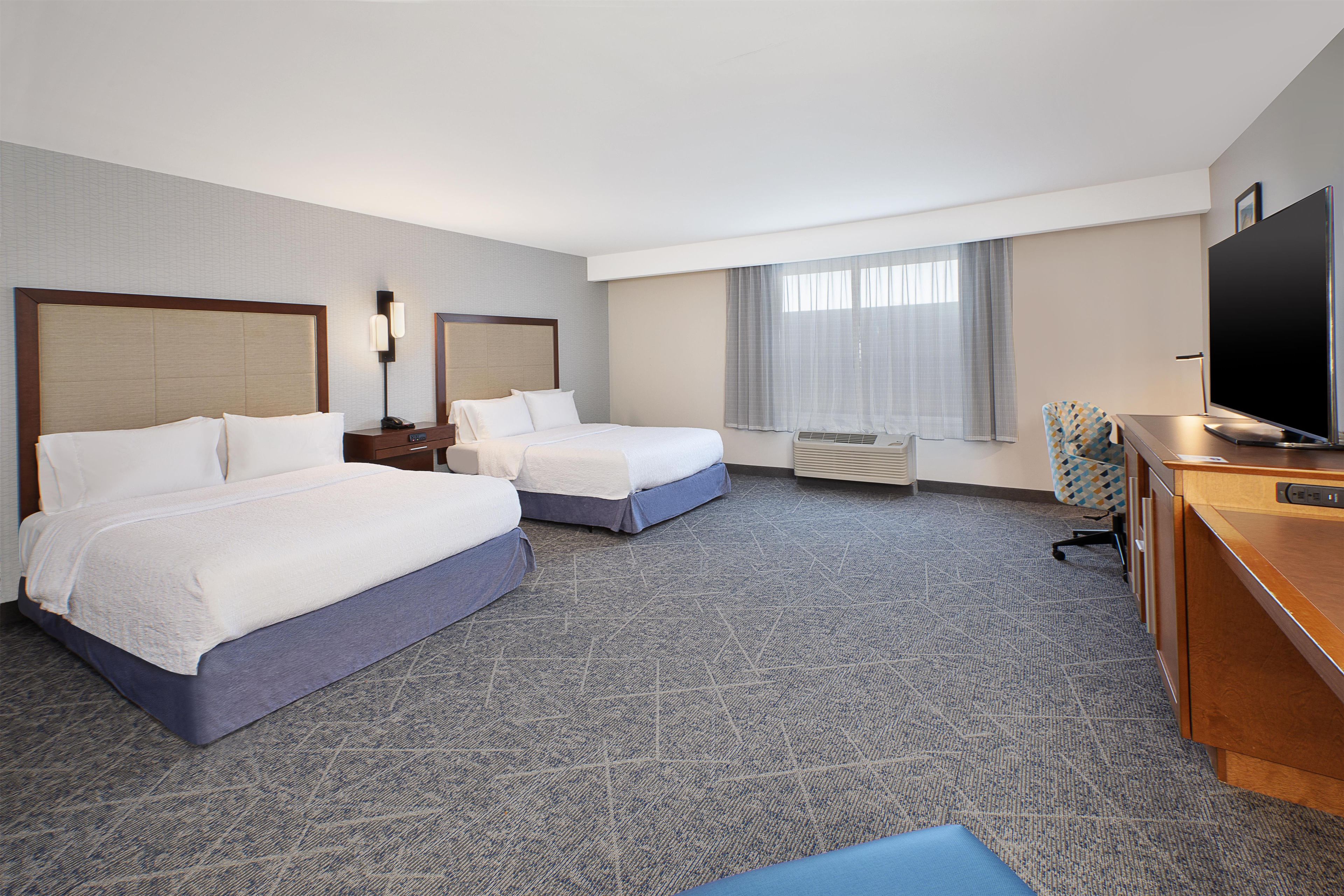 A hotel room with two beds, a desk, a television, and a large window with curtains.
