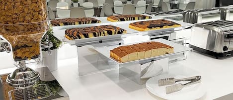 Disposición de bufé con diversos postres en bandejas, un dispensador de café y un tostador.