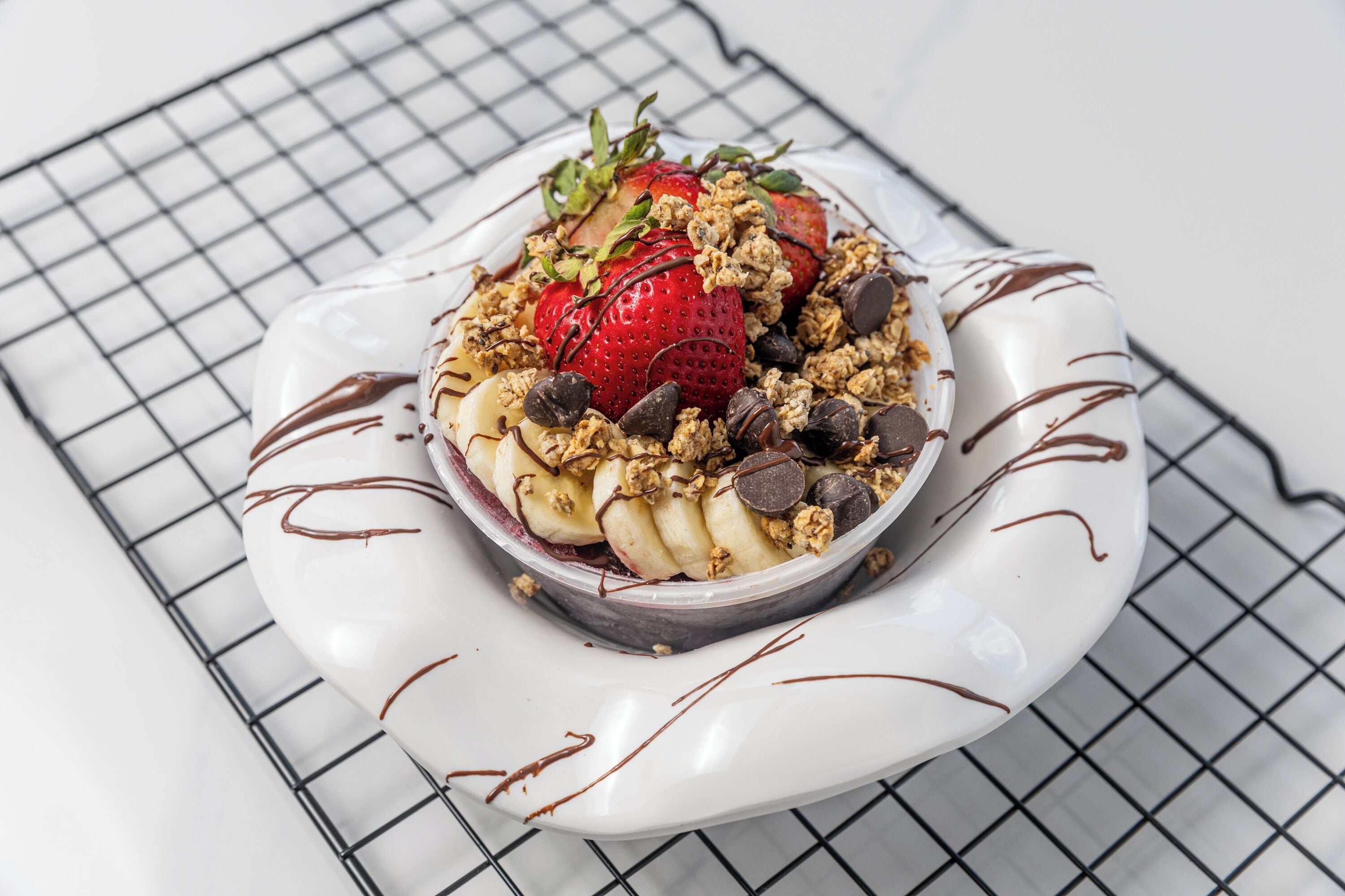 A dessert bowl on a white plate with chocolate drizzle, strawberries, banana slices, and granola.