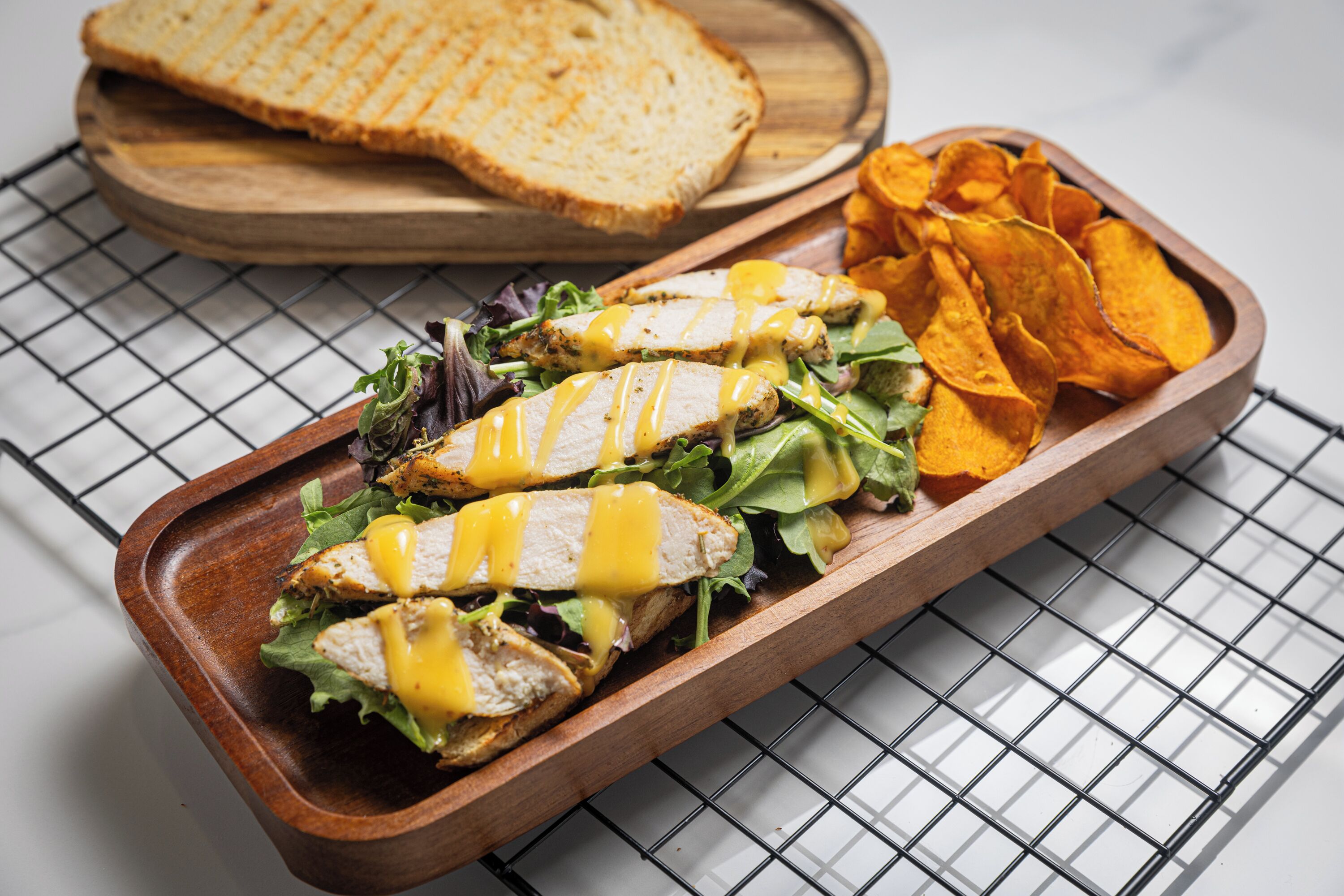 Een houten dienblad met een sandwich, chips en een stuk brood op een metalen rek.