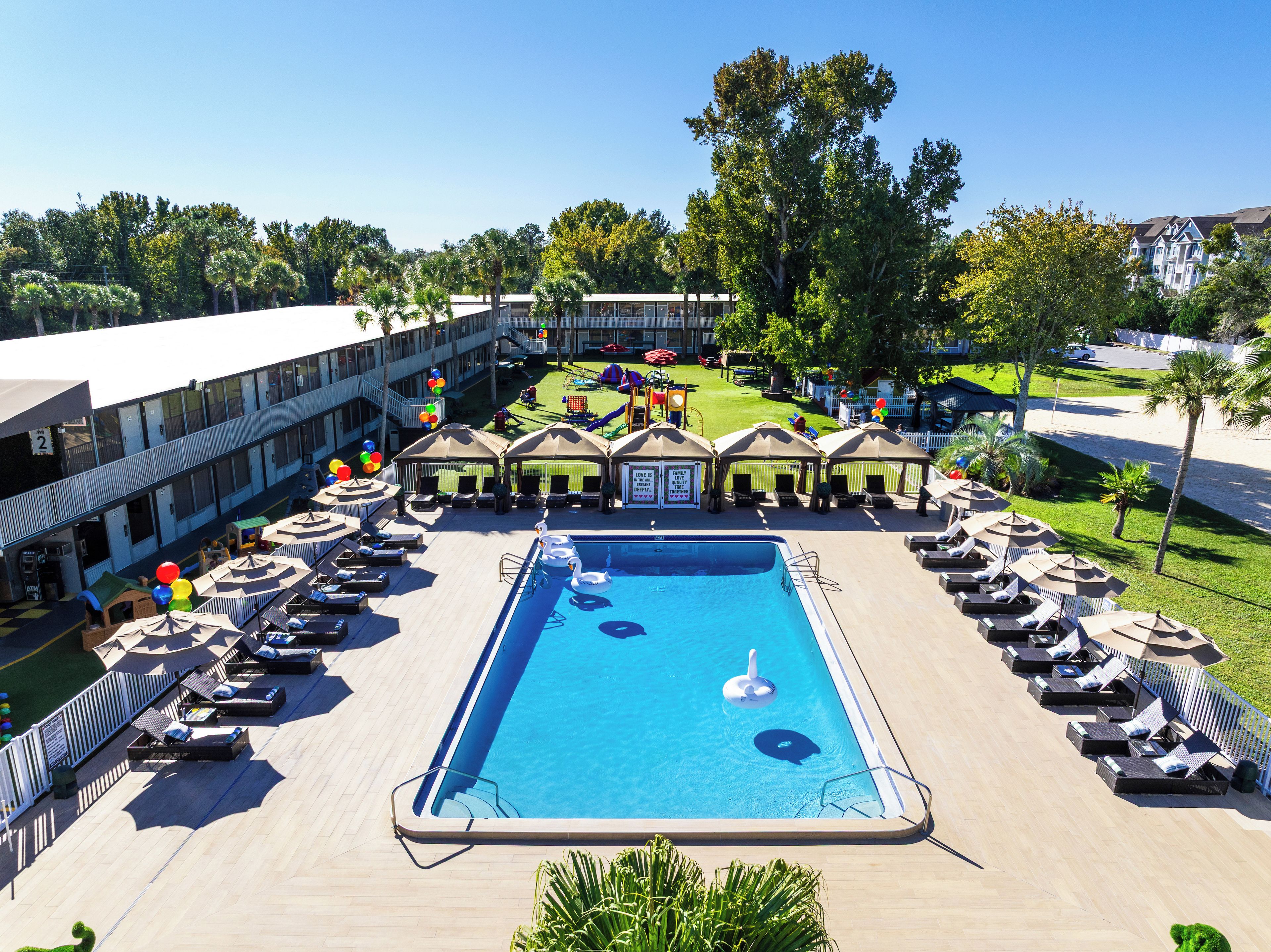 Zona de piscina con sillones de descanso y sombrillas, rodeada por un edificio de varios pisos y un jardín cercado.