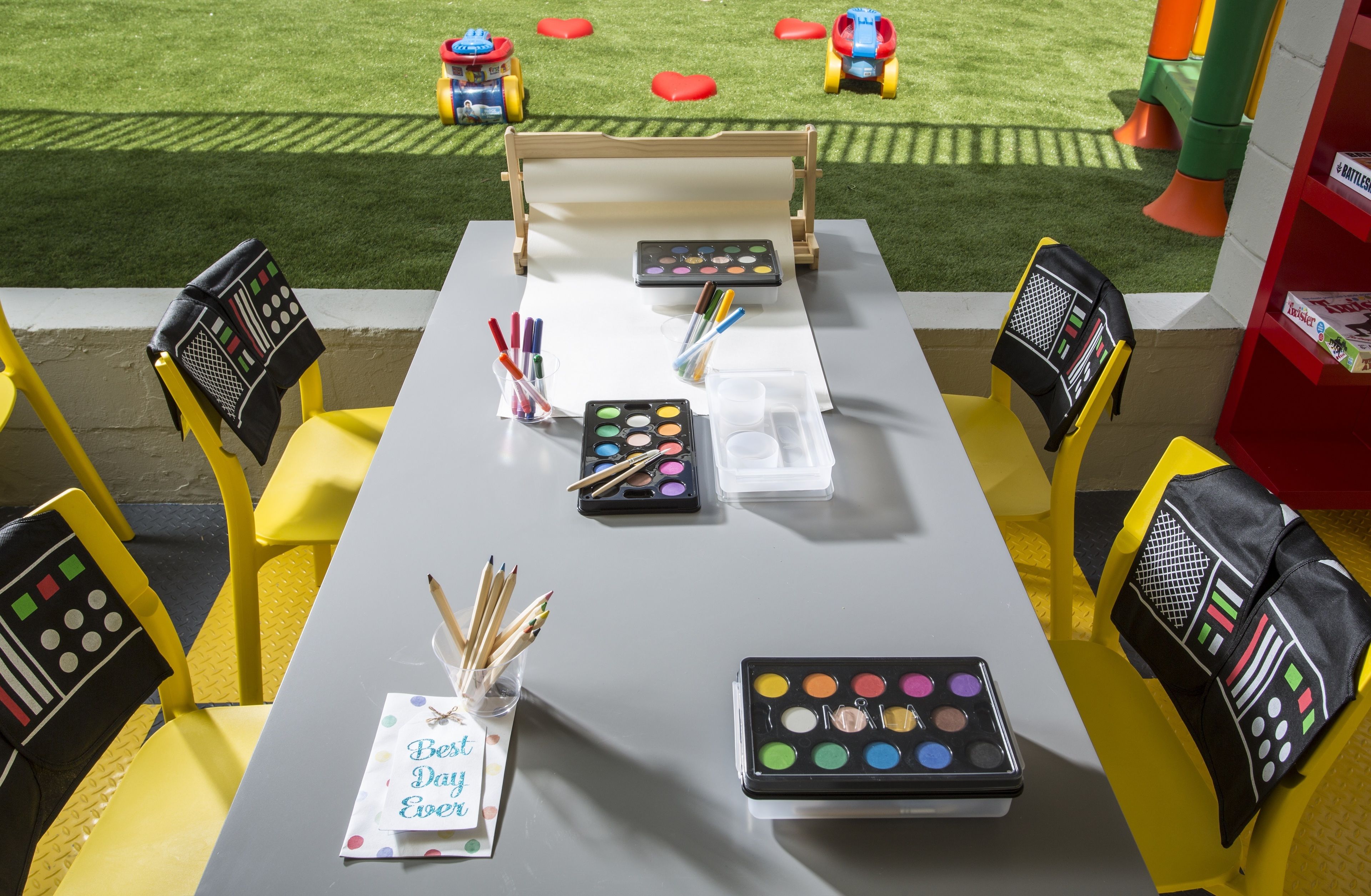 A table set up with art supplies, including paint palettes and brushes, in an outdoor area with a grassy lawn and children's toys in the background.