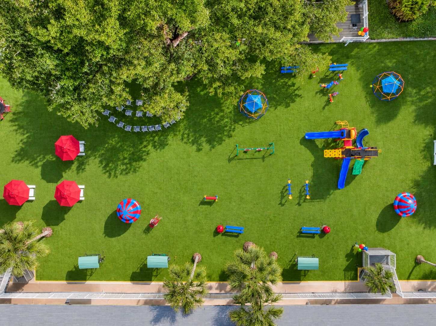 An aerial view of a playground with various play structures, including swings, slides, and climbing frames, surrounded by green grass and trees.