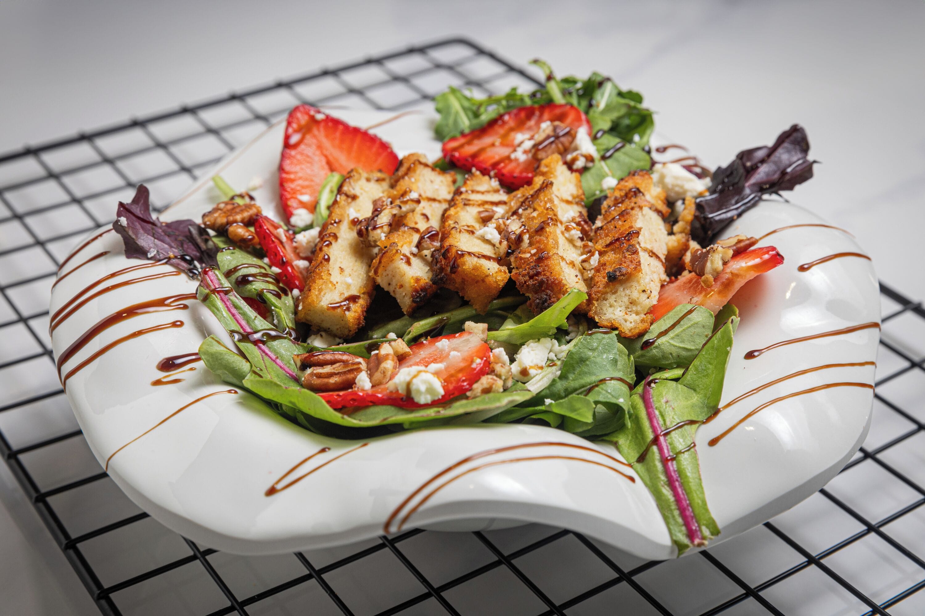 A white plate with a salad containing grilled chicken, strawberries, and greens, drizzled with a brown sauce.