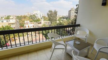 Un balcón con mobiliario de mimbre blanco, una pequeña mesa y vistas a un paisaje urbano con edificios y árboles.