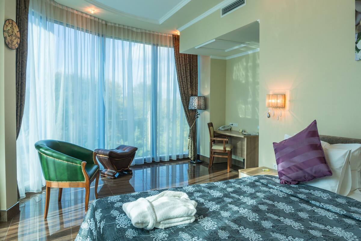 Quarto de hotel com cama, escrivaninha, cadeira e uma janela grande com cortinas.