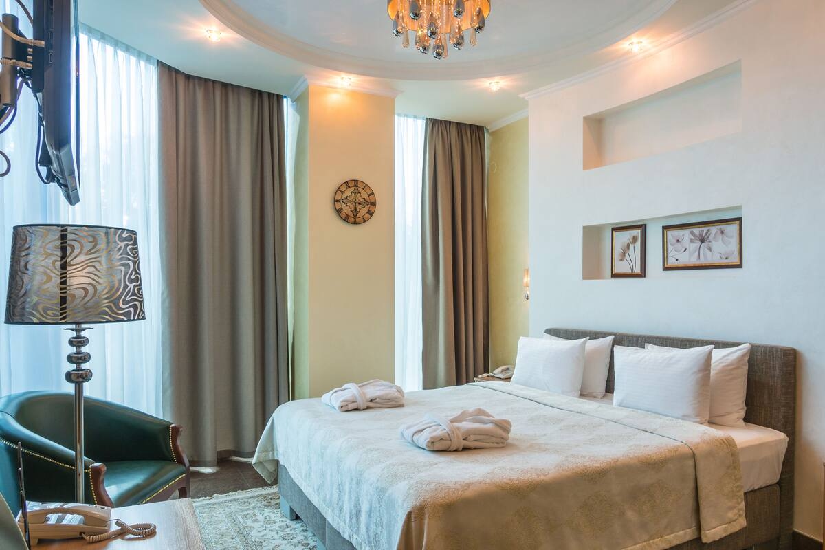 Quarto de hotel com cama, escrivaninha com telefone, cadeira, abajur e lustre.