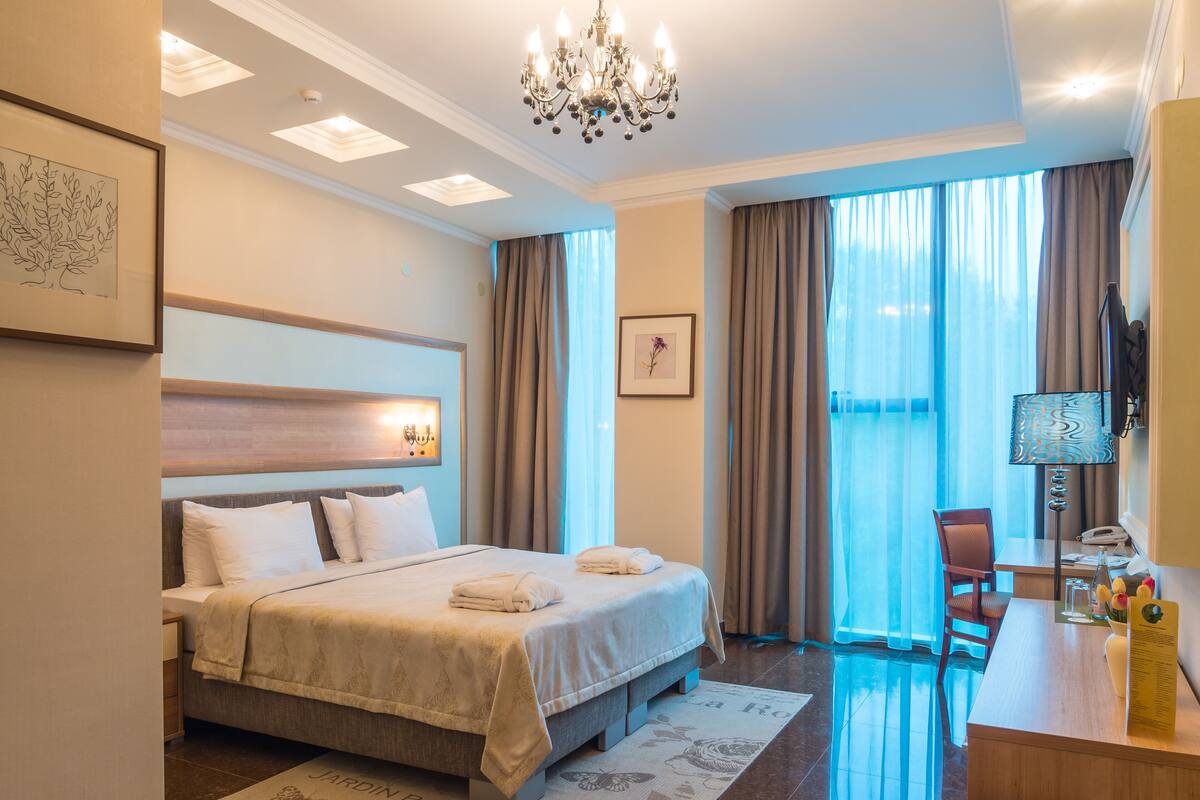 Quarto de hotel com uma cama grande, escrivaninha, cadeira, lustre e vista para o exterior pela janela.
