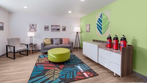 Un salon moderne avec un canapé, un pouf et un tapis coloré. On peut y voir des tableaux encadrés au mur, une lampe et un meuble contenant des thermos rouges.
