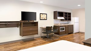Une chambre d’hôtel moderne avec un plancher en bois, un téléviseur à écran plat fixé au mur, une table à manger avec des chaises, un coin cuisine équipé d’un réfrigérateur et de placards.