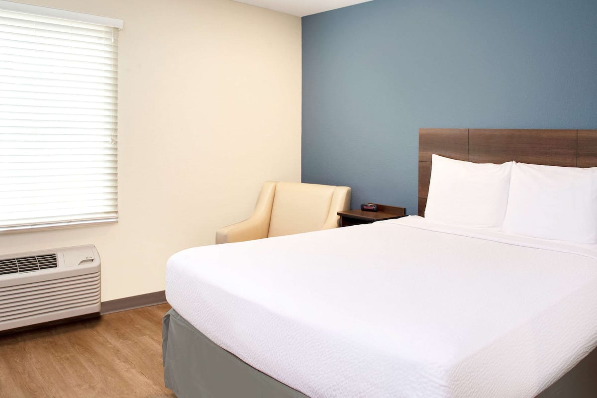Une chambre d’hôtel avec un grand lit, une chaise, une table de chevet et une fenêtre avec des stores.