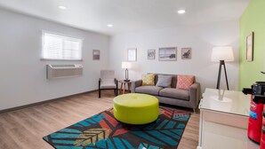 Un salon moderne avec un canapé, un pouf et une table basse. On y trouve une fenêtre avec des stores, une lampe et une petite table d’appoint.