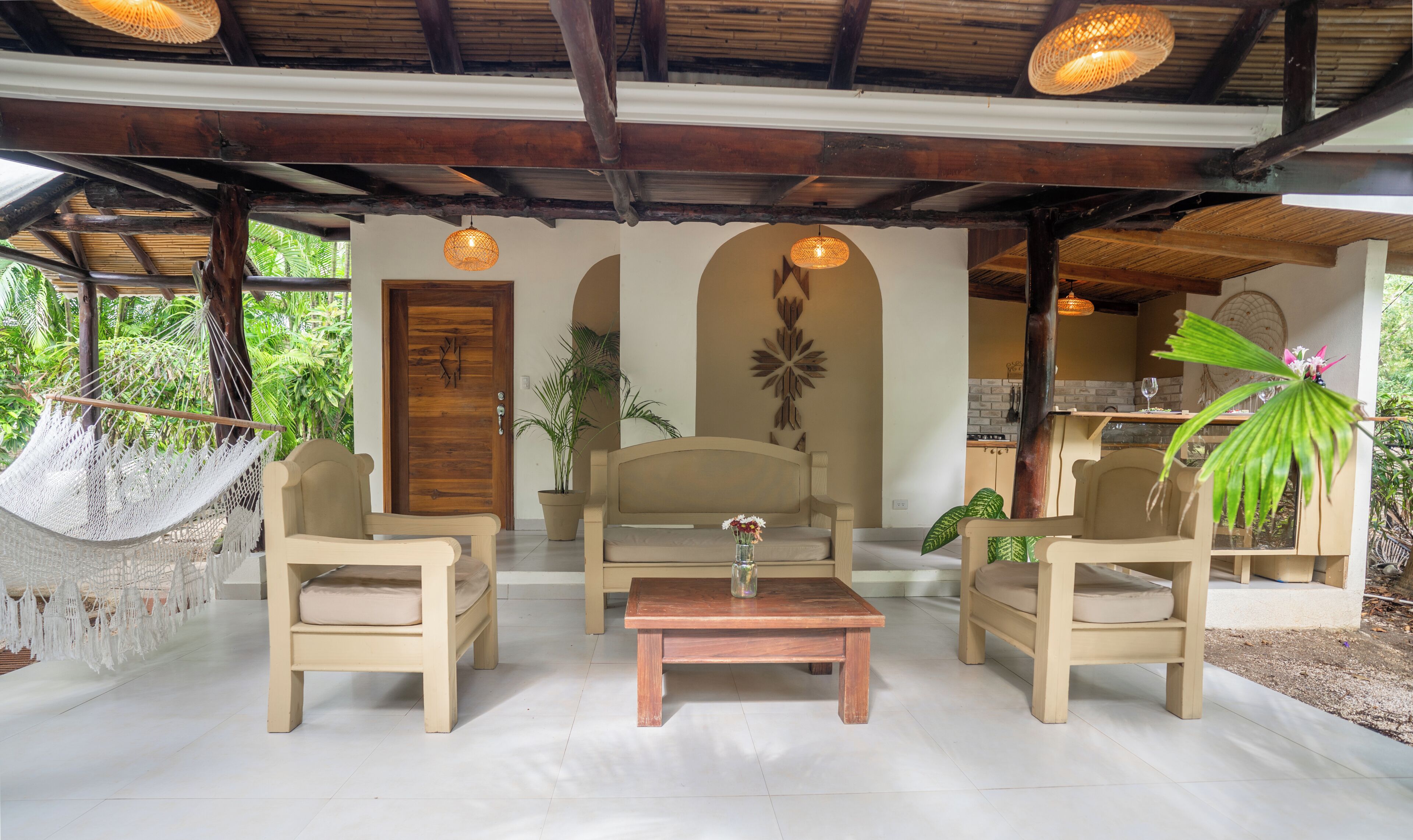 A room with a wooden door, a hammock, two beige chairs, a wooden table, and a potted plant.