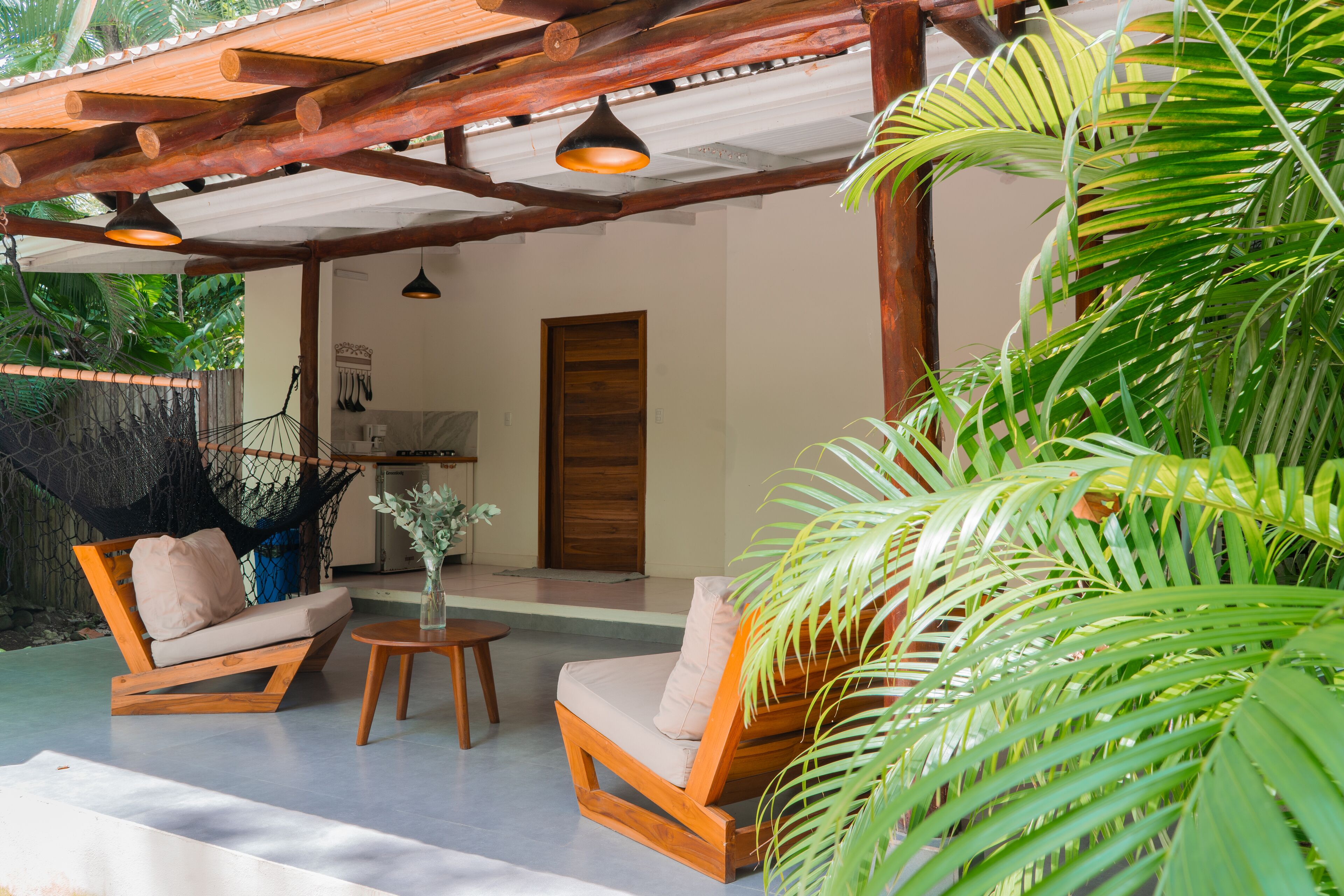 Een houten terras met twee comfortabele stoelen en een kleine tafel, omgeven door weelderig groen en een houten pergola met hangende lampjes.