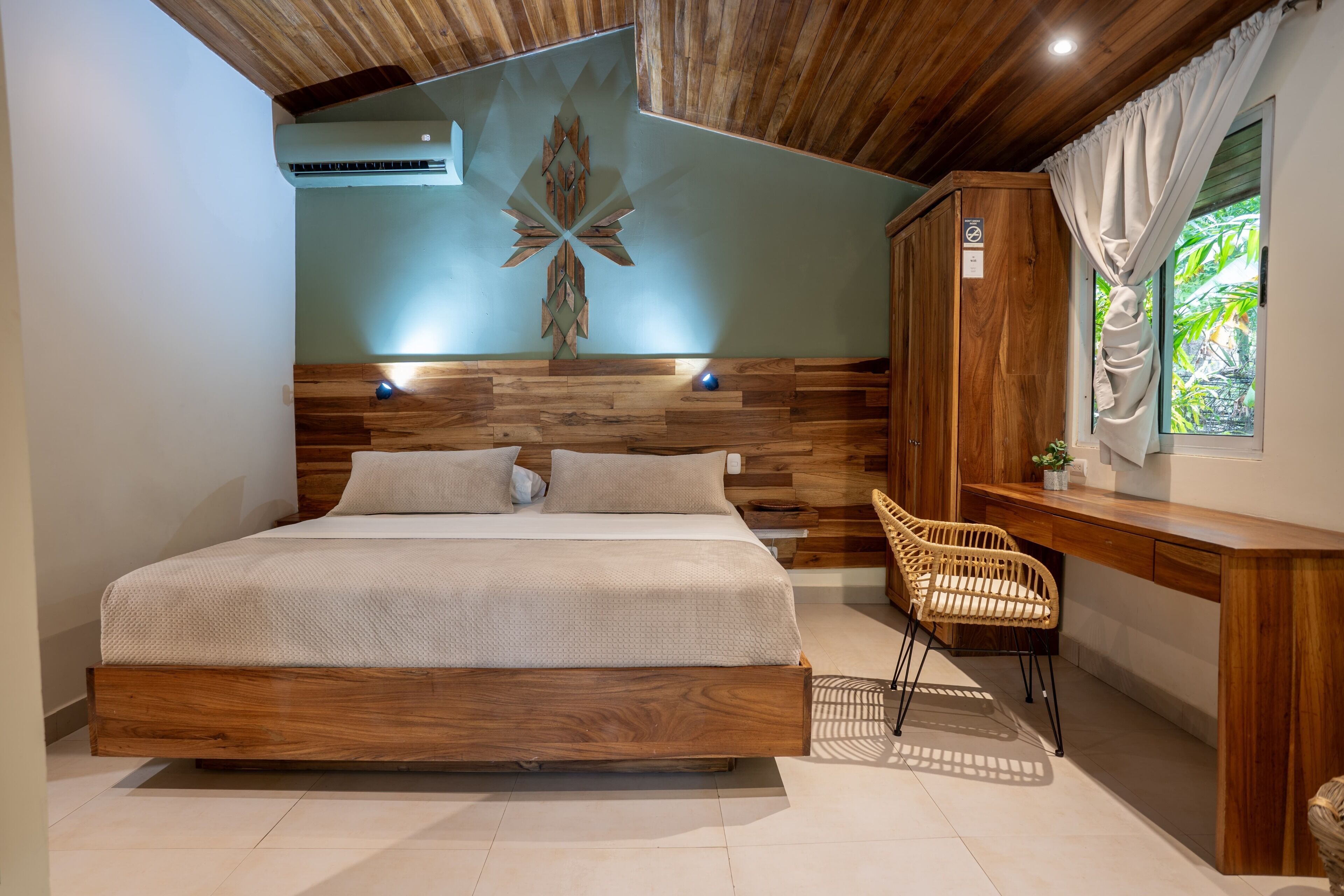 A bedroom with a wooden headboard, a bed with white bedding, a wicker chair, a wooden desk, and a window with curtains.