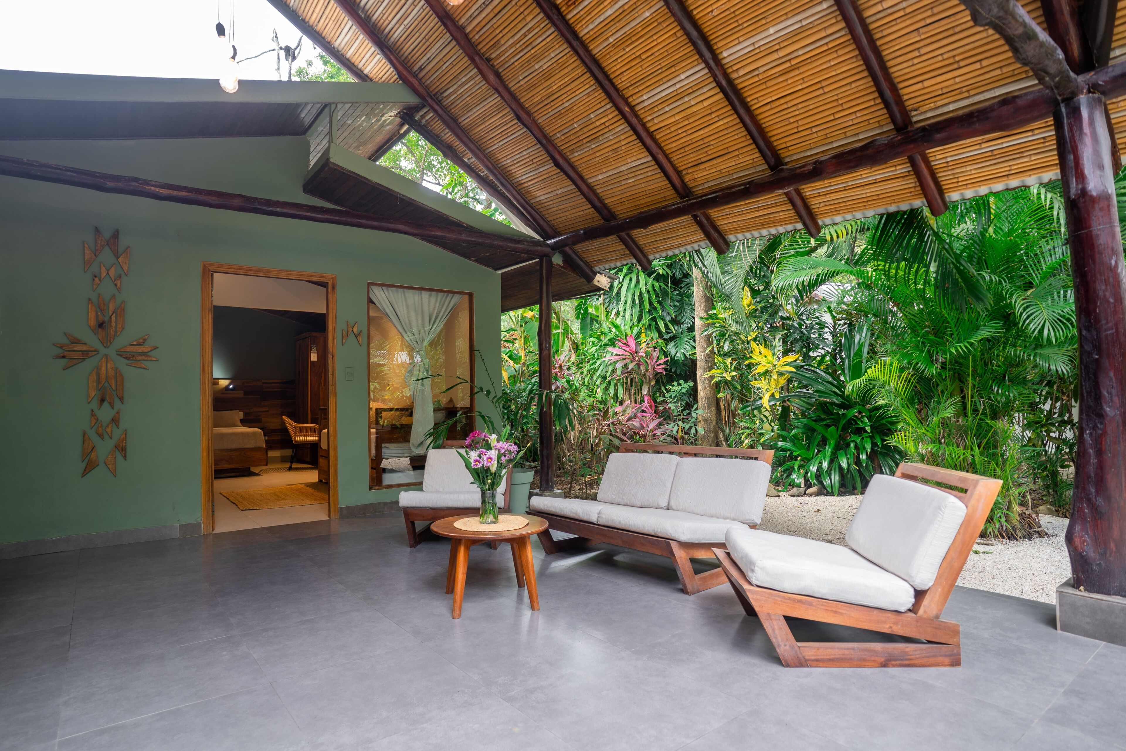 A covered outdoor seating area with a sofa and chairs, a small round table, and a view of lush greenery.