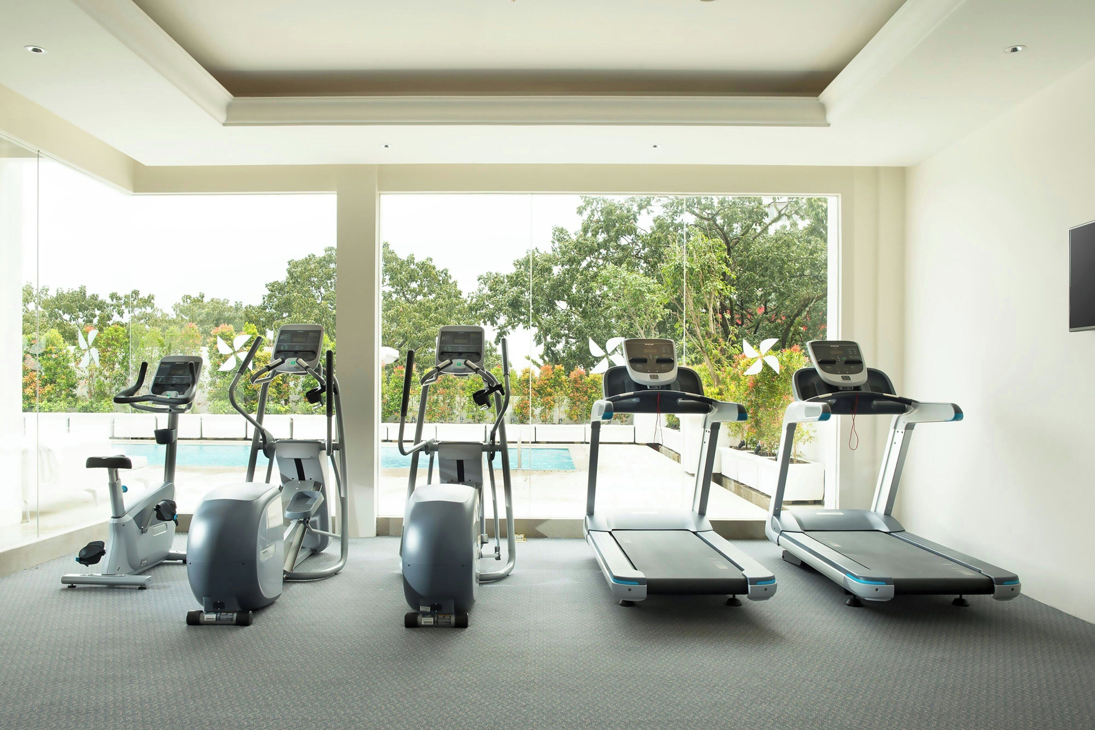 Ein Fitnessstudio mit vier Laufbändern und zwei Crosstrainern, große Fenster mit Blick auf Bäume und einen Pool.