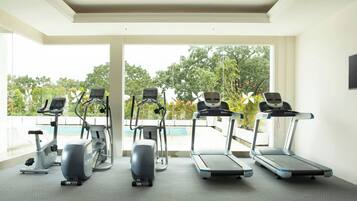 Academia com quatro esteiras e duas máquinas elípticas, janelas amplas com vista para árvores e uma piscina.