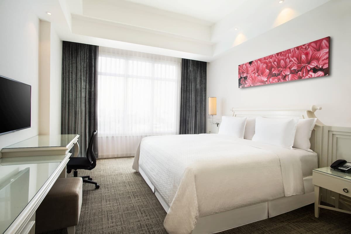 Una habitación de hotel con una cama grande, un escritorio con silla, un televisor, una mesita de noche con lámpara y un cuadro en la pared.