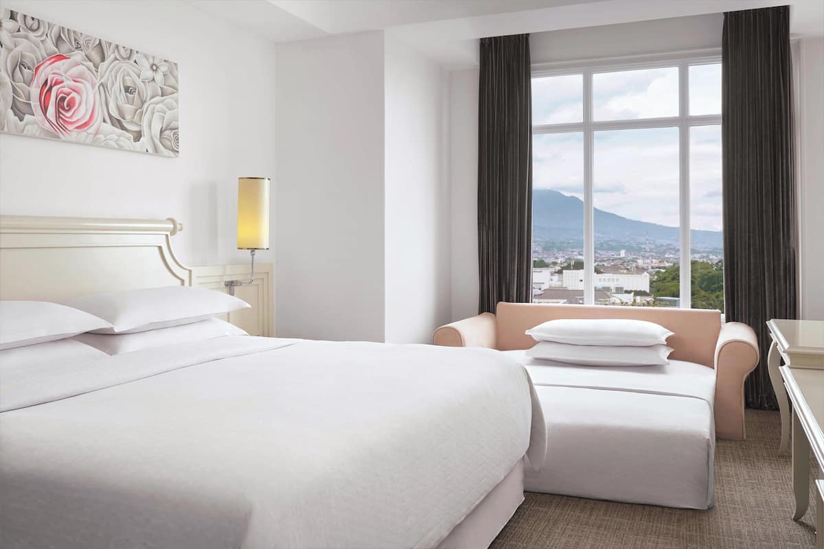 Habitación de hotel con una cama grande, un sofá, un escritorio, una lámpara y una ventana con vistas a la ciudad.