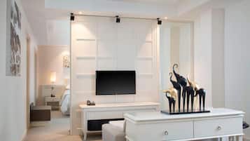 A hotel room with a large flat-screen TV mounted on a white wall, a white console table with drawers, a sofa, and a small bedside table with a lamp.