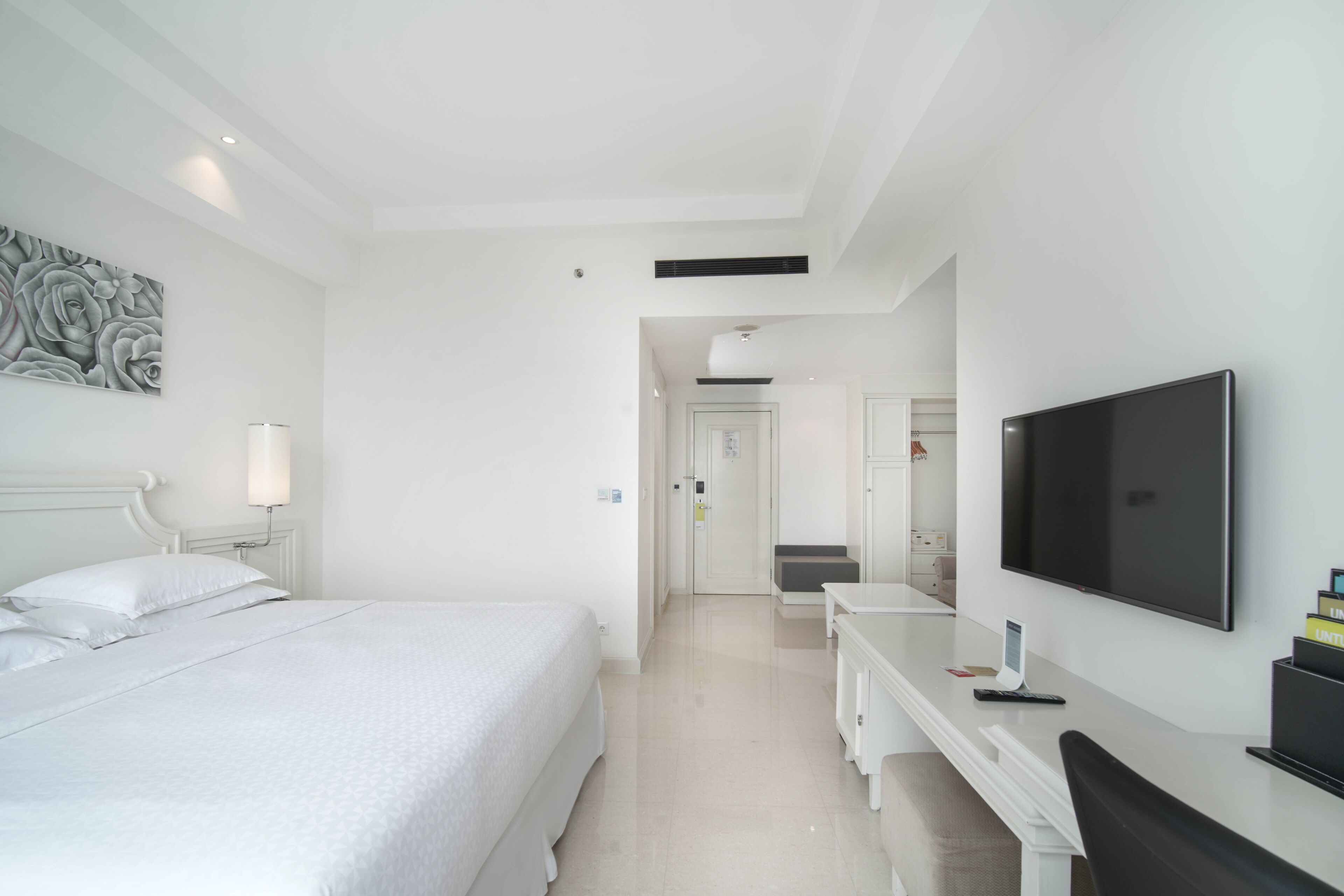 A hotel room with a large bed, a desk with a flat-screen TV, a bedside lamp, and a wall-mounted artwork.