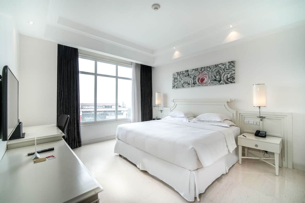 Una habitación de hotel con una cama grande, dos mesitas de noche con lámparas, un escritorio con una silla y una ventana con cortinas.