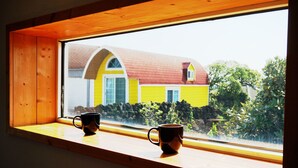 Une fenêtre avec deux tasses noires posées sur un rebord en bois, donnant sur une maison jaune avec un toit rouge et un mur en pierre.
