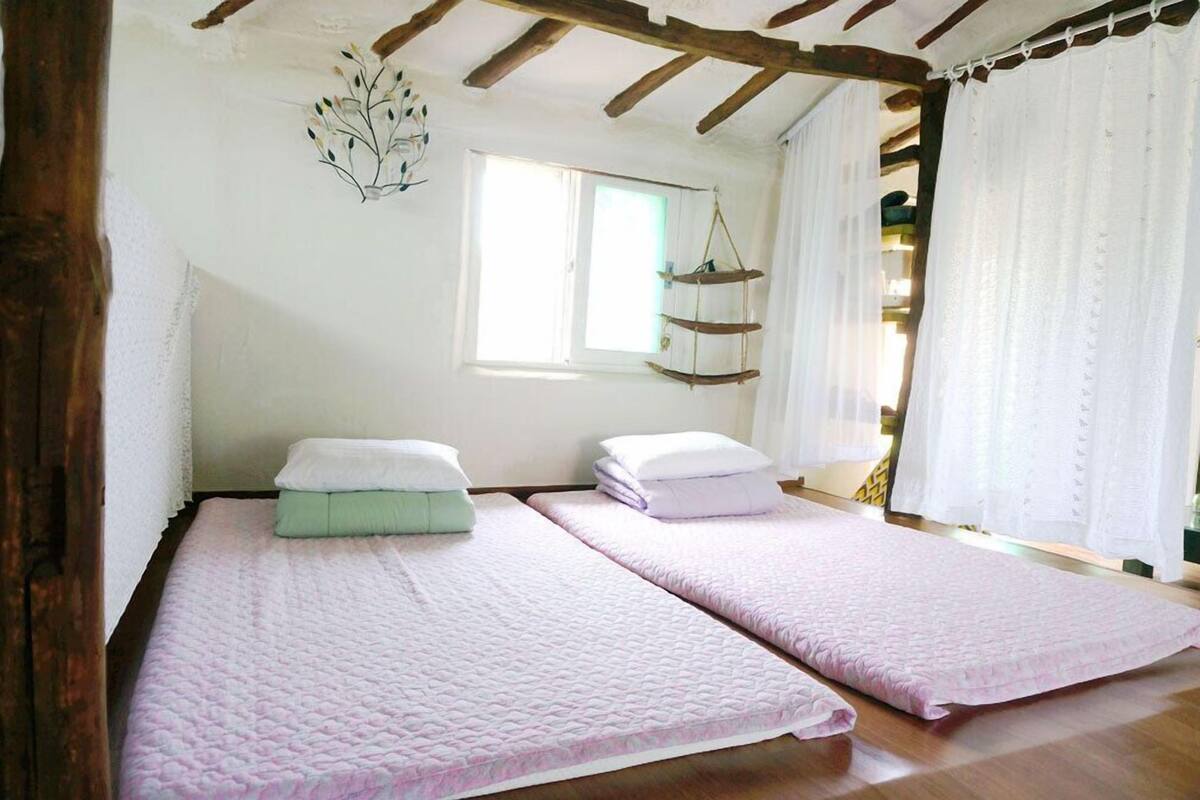 A room with two beds, wooden beams, a hanging plant, and a window with curtains.