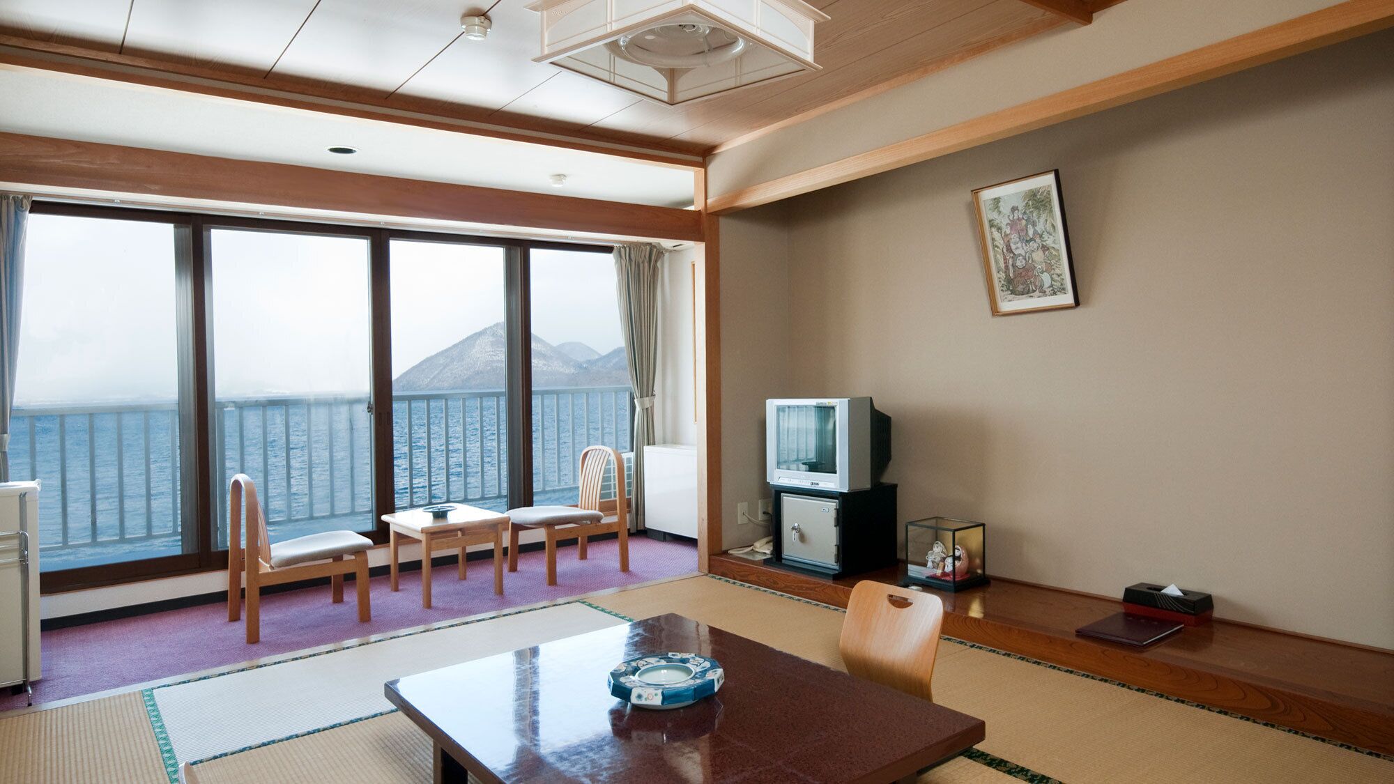 A room with a large window offering a view of the sea and mountains, a television, a small cabinet, and a table with a decorative item.
