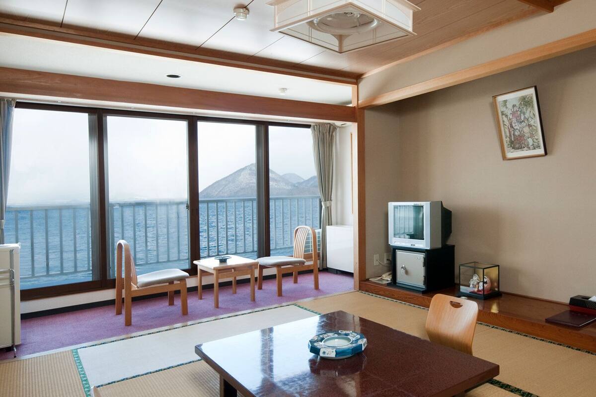 A room with a large window offering a view of the sea and mountains, a television, a small cabinet, and a table with a decorative item.