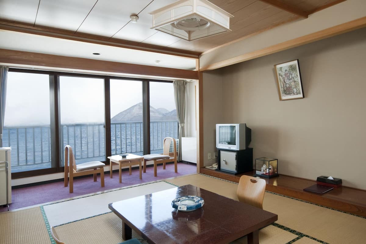 A room with a large window overlooking a body of water and mountains, a wooden table, a television, and a framed picture on the wall.