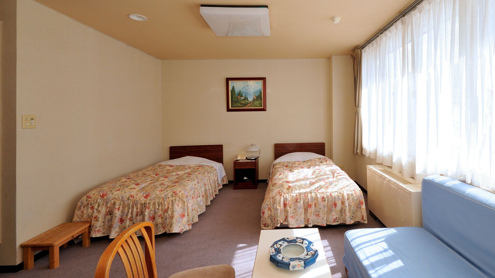 A room with two beds, a small table, a chair, and a window with curtains.
