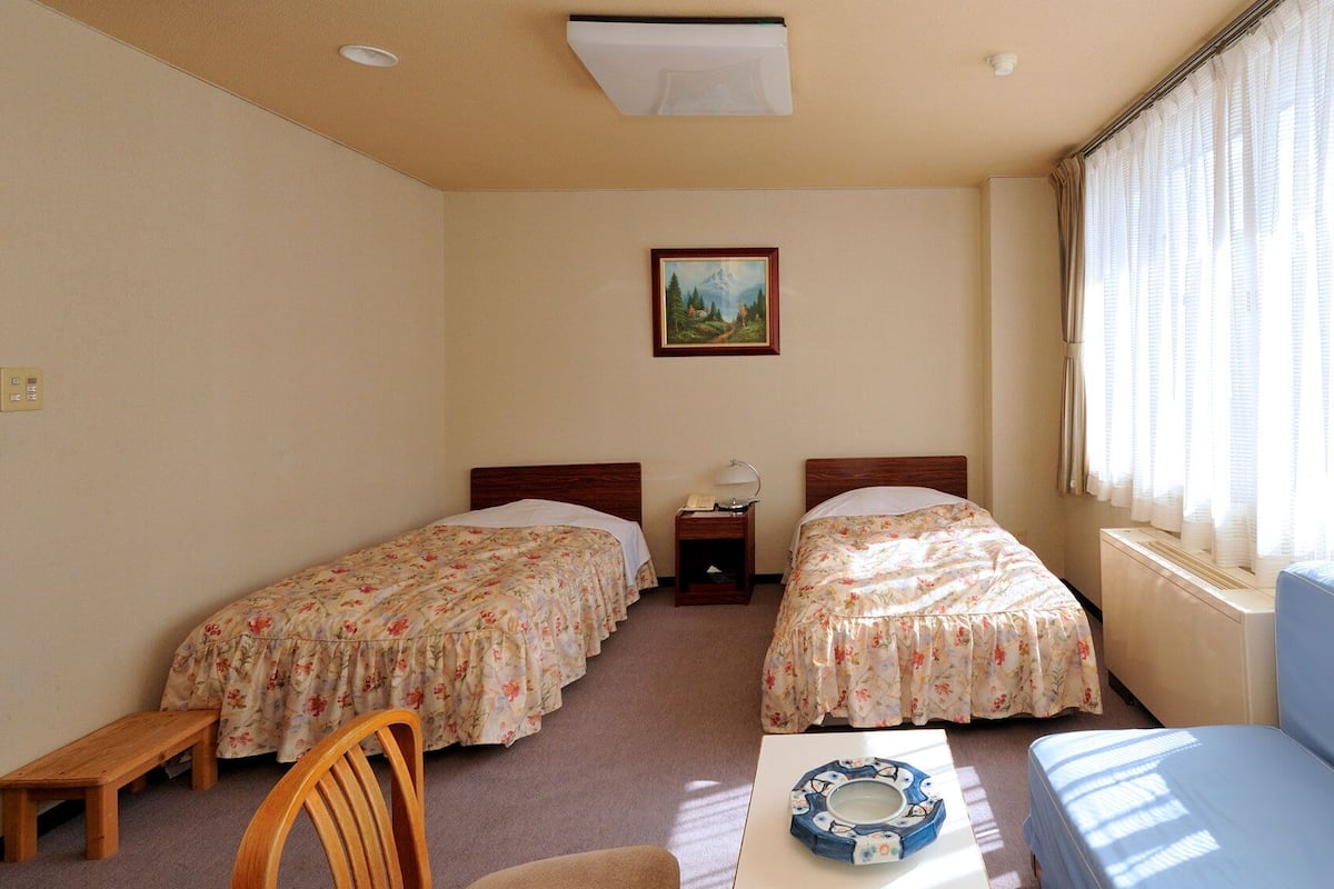 Quarto com duas camas, uma mesa pequena, uma cadeira e janela com cortinas.