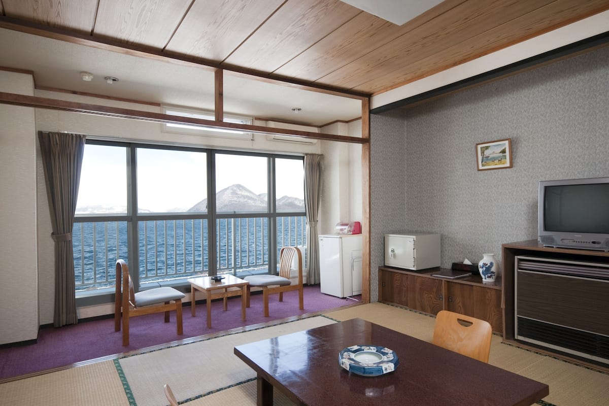 A room with a large window overlooking a body of water, a dining table, chairs, a television, and a small refrigerator.