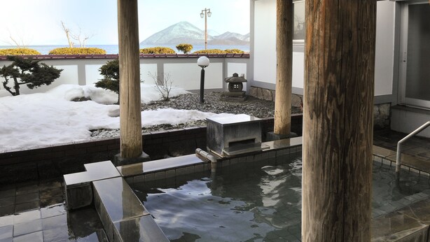 Un onsen japonais traditionnel, avec une source d’eau chaude naturelle alimentée par des pierres, des piliers en bois et une vue sur des montagnes enneigées.