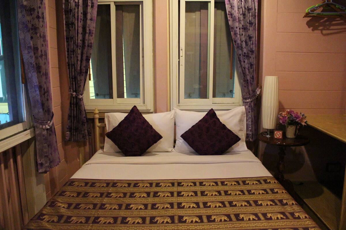 A bedroom with two windows, a bed with patterned bedding, and a nightstand with a lamp and flowers.