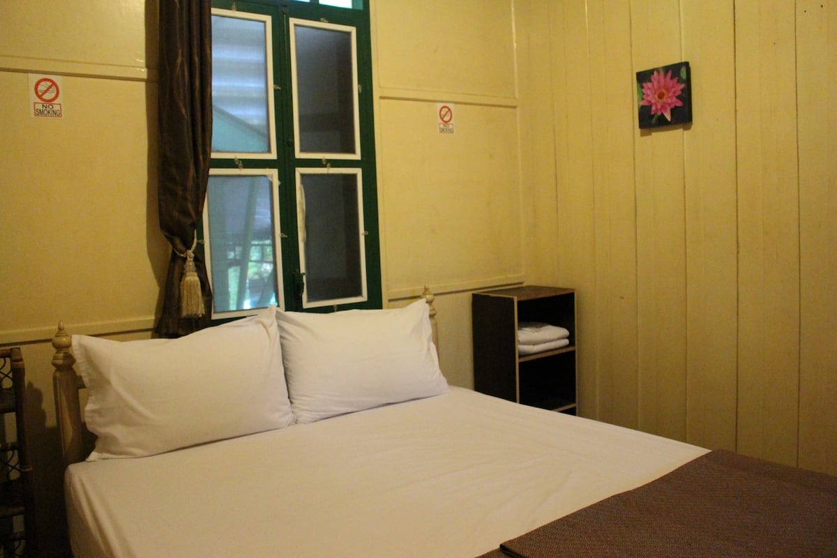 A bed with white linens, a wooden headboard, a small shelf with folded towels, and a window with curtains.